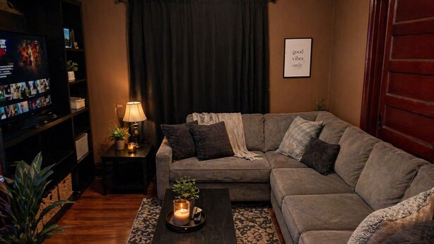 A living room with a sectional sofa, a coffee table with a candle, a TV on a stand, and a wall with a framed sign.