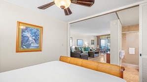 Ein Schlafzimmer mit einem Bett, einem Deckenventilator, einem gerahmten Bild und Blick in einen Wohnbereich mit Möbeln.