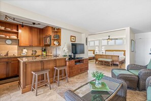 A modern kitchen with wooden cabinets, a central island, bar stools, a flat-screen TV, and a dining area with wicker furniture. - New Listing! Oceanfront Island Escape at Hale Ono Loa 207 (Lahaina)