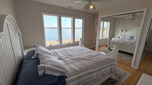 Une chambre à coucher avec un lit, une grande fenêtre donnant sur l’eau, un ventilateur de plafond et un miroir.