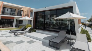 Een modern terras buiten met ligbedden en parasols, een zwembad en een gebouw met een glazen gevel.