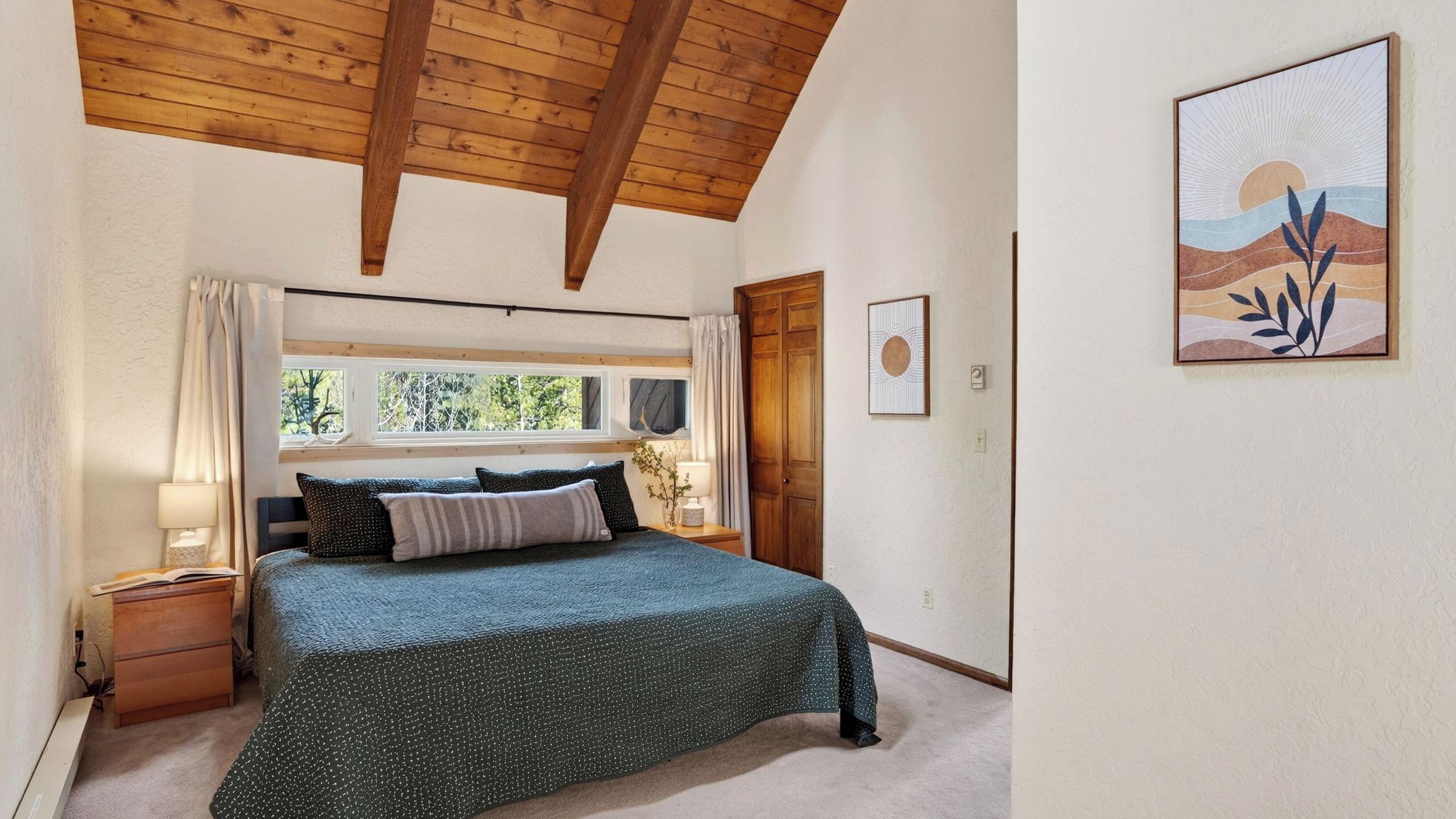 Un dormitorio con cama, vigas de madera en el techo, una ventana con cortinas, una mesita de noche con lámpara y un cuadro en la pared.
