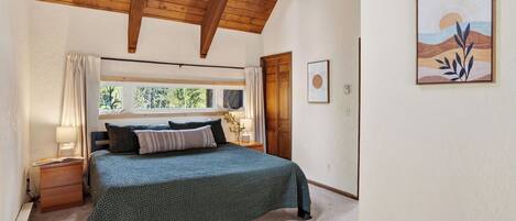 Un dormitorio con cama, vigas de madera en el techo, una ventana con cortinas, una mesita de noche con lámpara y un cuadro en la pared.