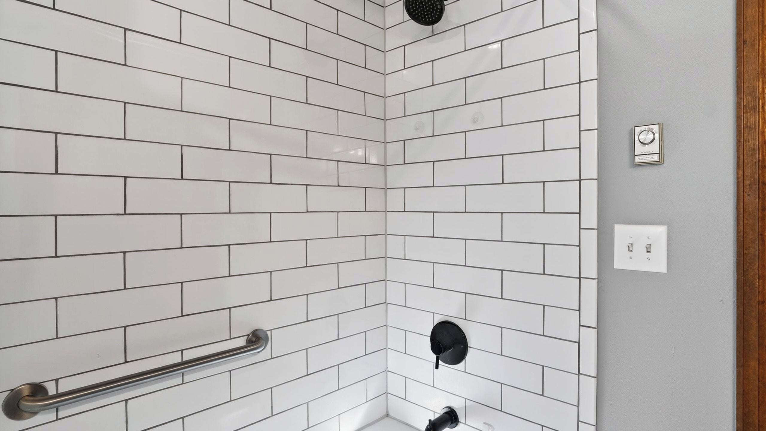 A bathroom with white subway tiles, a handrail, a showerhead, and a thermostat on the wall.