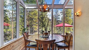 Une table en bois entourée de chaises, un lustre et un parasol rouge dans une pièce dotée de grandes fenêtres et offrant une vue sur les arbres.