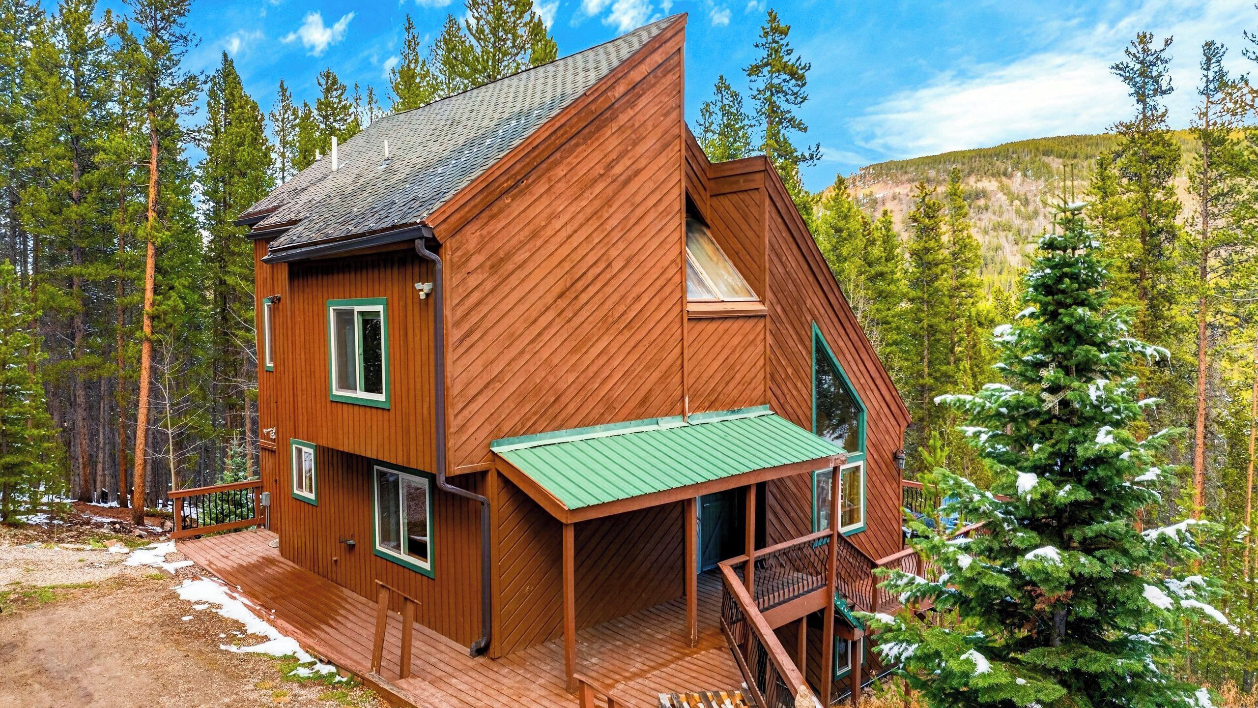 A wooden cabin with a green roof, a balcony, and a view of a forested mountain.