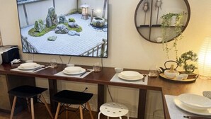 Ein Essbereich mit einem langen Tisch, gedeckt für zwei Personen, einem Fernseher, der eine Gartenszene zeigt, und einem runden Spiegel an der Wand.