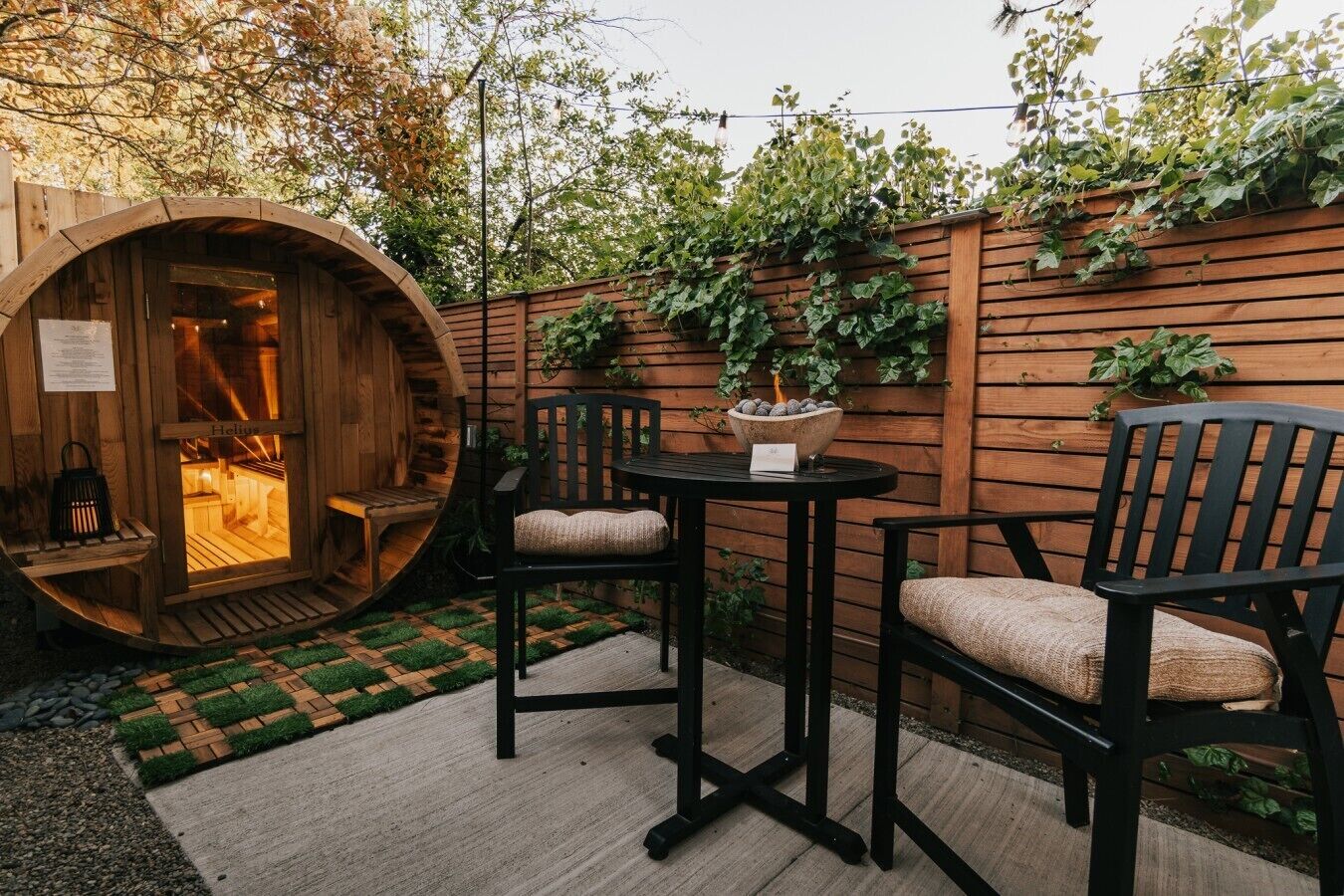 Un área de estar al aire libre con muebles de madera, una mesa redonda y dos sillas, una estructura de barril de madera con fuego en su interior, y una valla de madera cubierta de plantas trepadoras.