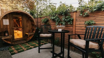 Un área de estar al aire libre con muebles de madera, una mesa redonda y dos sillas, una estructura de barril de madera con fuego en su interior, y una valla de madera cubierta de plantas trepadoras.