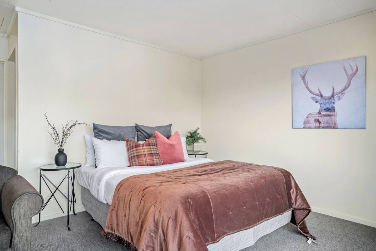 A bedroom with a bed, pillows, a side table with a vase, a chair, and a wall-mounted painting of an elk.