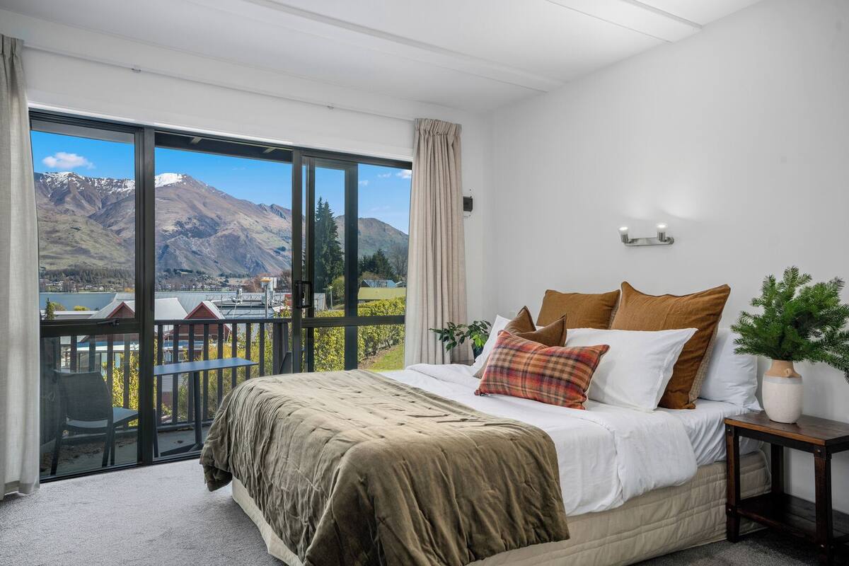A bedroom with a large bed, a nightstand, a potted plant, and a view of mountains through a balcony.