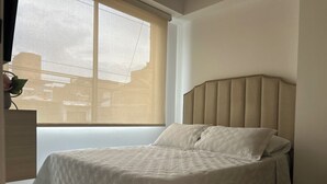 Una cama con cabecera, una ventana con persianas y una planta sobre un pedestal.