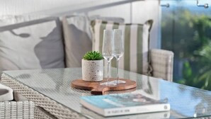 Een glazen tafel met een boek, een kleine plant in pot en twee glaasjes op een houten onderzetter.