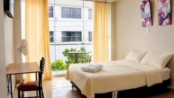 A hotel room with a bed, a desk, a chair, a window with curtains, and artwork on the wall.