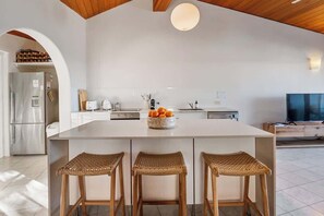 A modern kitchen with a large island, three wicker stools, a bowl of fruit, and a TV in the background.