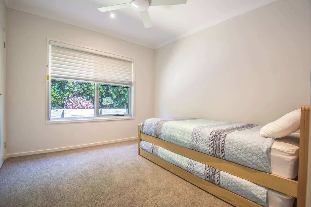 A room with a single bed, a window with blinds, a ceiling fan, and a carpeted floor.