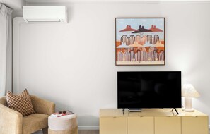 A living room with a sofa, a coffee table, a television on a stand, a painting on the wall, and an air conditioning unit. - Chic Noosa Riverside Apt with Great Facilities (Noosa Region)