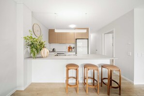 A modern kitchen with a central island, wooden stools, and a hanging light fixture. - Chic Noosa Riverside Apt with Great Facilities (Noosa Region)