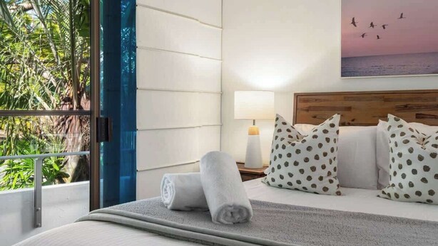A hotel room with a bed, pillows, a nightstand, a lamp, and a view of greenery and a sunset. - Resort Unit, Heated Pool, AC, Walk to Main Beach (Noosa Region)