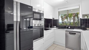 A modern kitchen with stainless steel appliances, white cabinetry, and a view of palm trees. - Resort Unit, Heated Pool, AC, Walk to Main Beach (Noosa Region)