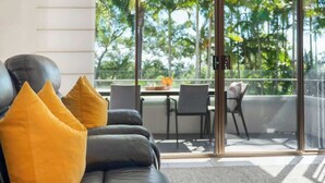 A modern living room with a black leather sofa, yellow cushions, and a view of a balcony with outdoor furniture. - Resort Unit, Heated Pool, AC, Walk to Main Beach (Noosa Region)