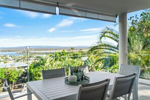 An outdoor patio with a table set for two, surrounded by chairs, overlooking a scenic view of a body of water and greenery. - Noosa Hill Apt, Views, Pool, with Hasting St, Beach (Noosa Region)