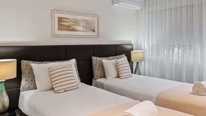 Ett hotellrum med två sängar, en inramad tavla på väggen, en luftkonditionering och ett fönster med tunna, genomskinliga gardiner.
