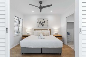 A modern bedroom with a large bed, a ceiling fan, bedside lamps, and a wall-mounted abstract art piece. - Montpellier Riverside Apartment with Heated Pool (Noosa Region)