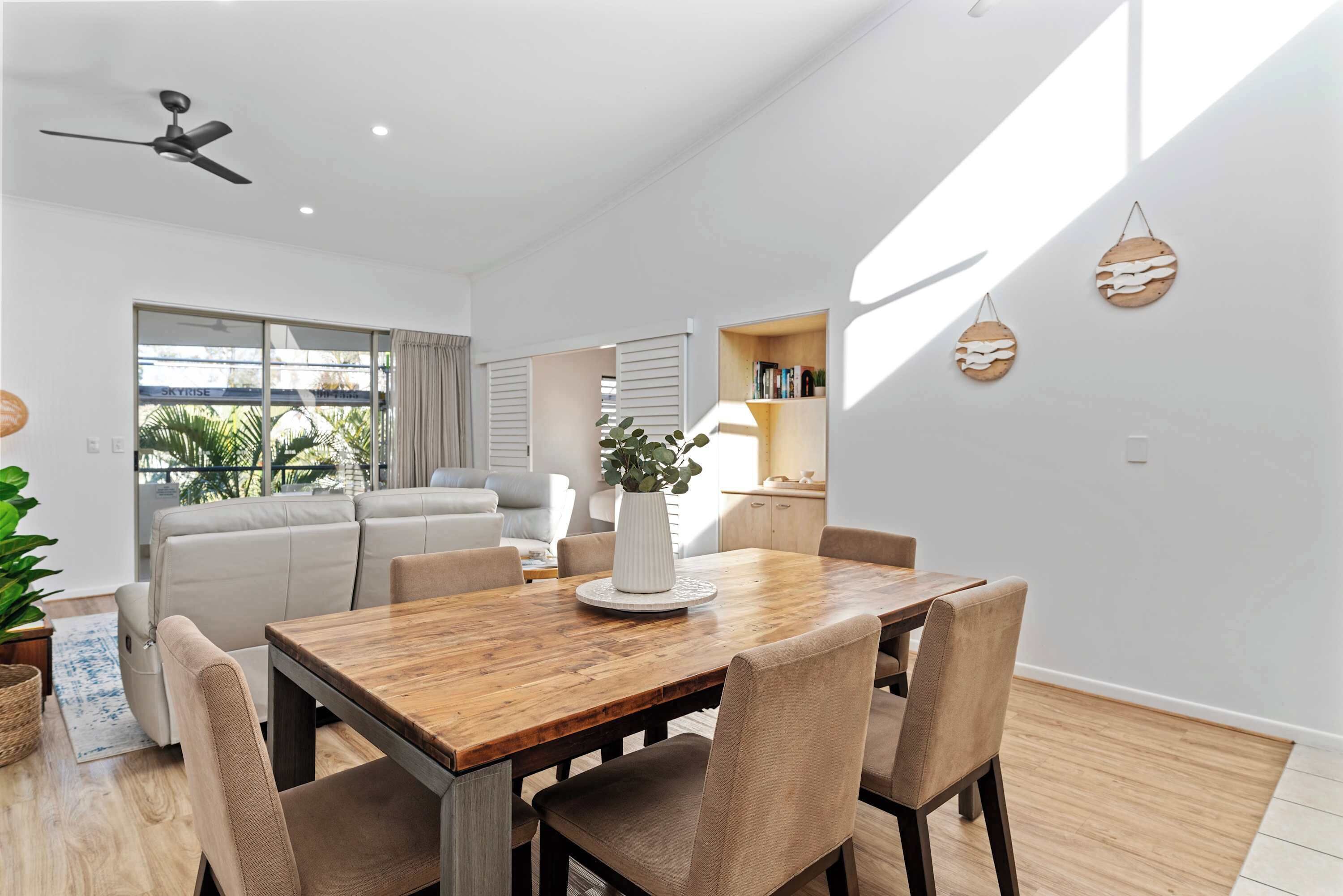 Ein moderner Essbereich mit Holztisch und -stühlen, einem Deckenventilator und Blick in ein Wohnzimmer mit Sofa und Pflanze.