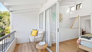 En balkong med ett vitt räcke, en flätad stol med kuddar, ett litet runt bord och en säng med ett randigt täcke.