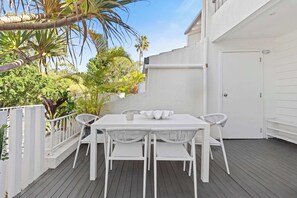 Une terrasse extérieure blanche, avec une table et des chaises, entourée d’une clôture blanche et d’une végétation luxuriante.