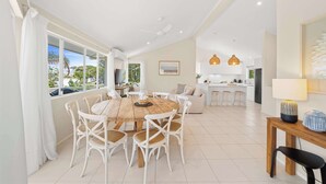 Amplia sala de estar con una mesa de comedor, sillas de mimbre blancas, una cocina al fondo y ventanas grandes.