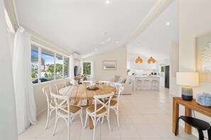 A spacious living area with a dining table, white wicker chairs, a kitchen in the background, and large windows. - Southwind in Sunrise (Noosa Region)
