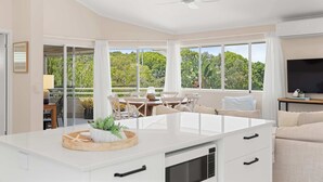 Una cocina moderna con una isla central, ventanas grandes con cortinas, un comedor con sillas y un televisor sobre un mueble.