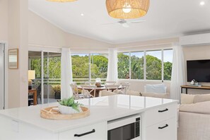 A modern kitchen with a central island, large windows with curtains, a dining area with chairs, and a TV on a stand. - Southwind in Sunrise (Noosa Region)