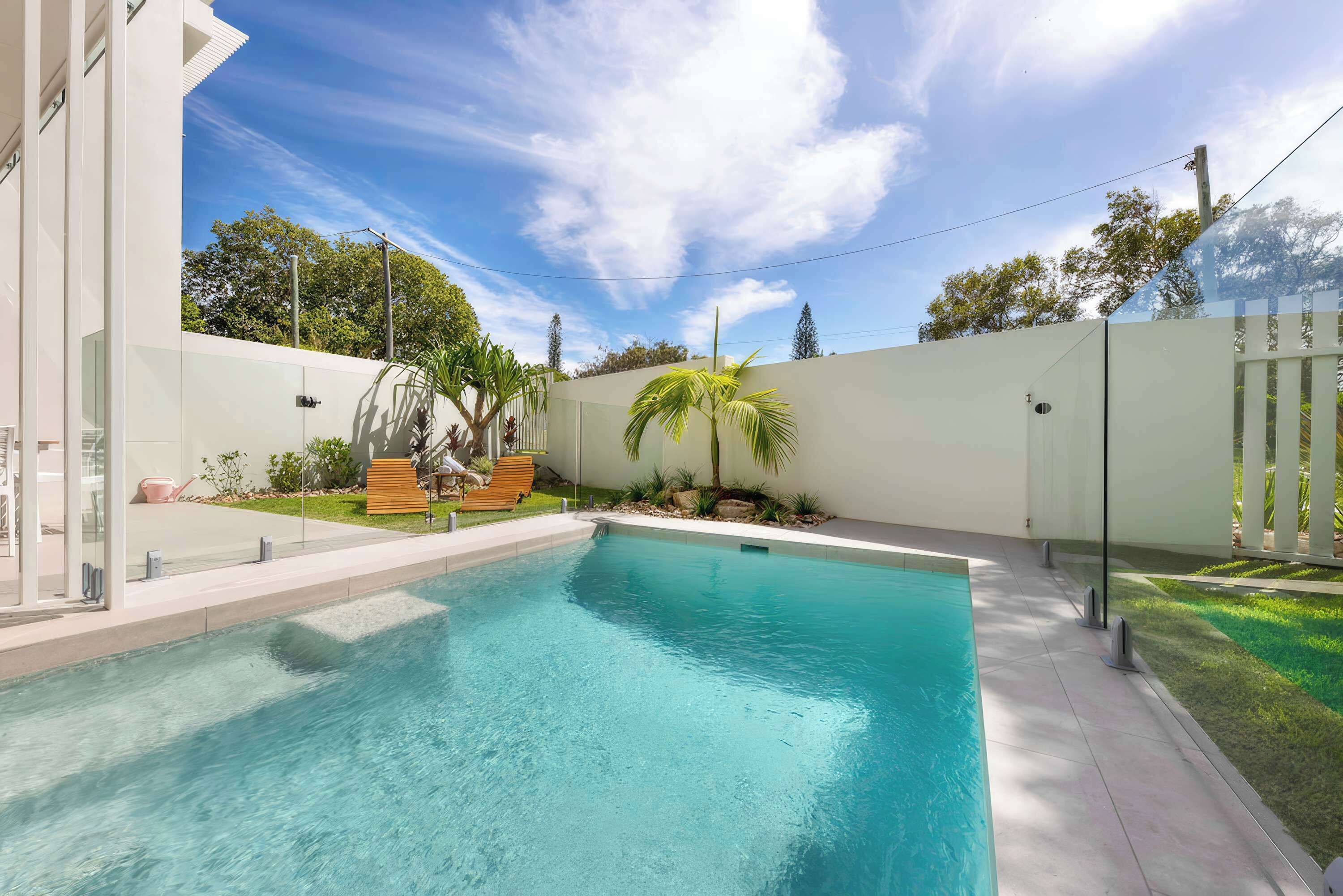 A swimming pool with a clear blue water surface, surrounded by a white wall and a grassy area with a wooden deck and lounge chairs.