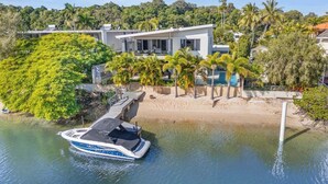 Uma casa moderna com piscina, rodeada por palmeiras e um barco atracado na água.