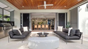 Sala de estar moderna com um banquinho redondo branco, dois sofás cinzas, um ventilador de teto e uma cozinha visível através de portas de vidro.