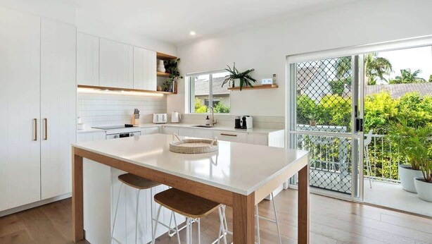 A modern kitchen with white cabinetry, a central island with stools, and a view of a green outdoor area through a glass door. - Modern, Stylish, Resort style Noosa River stay (Noosa Region)
