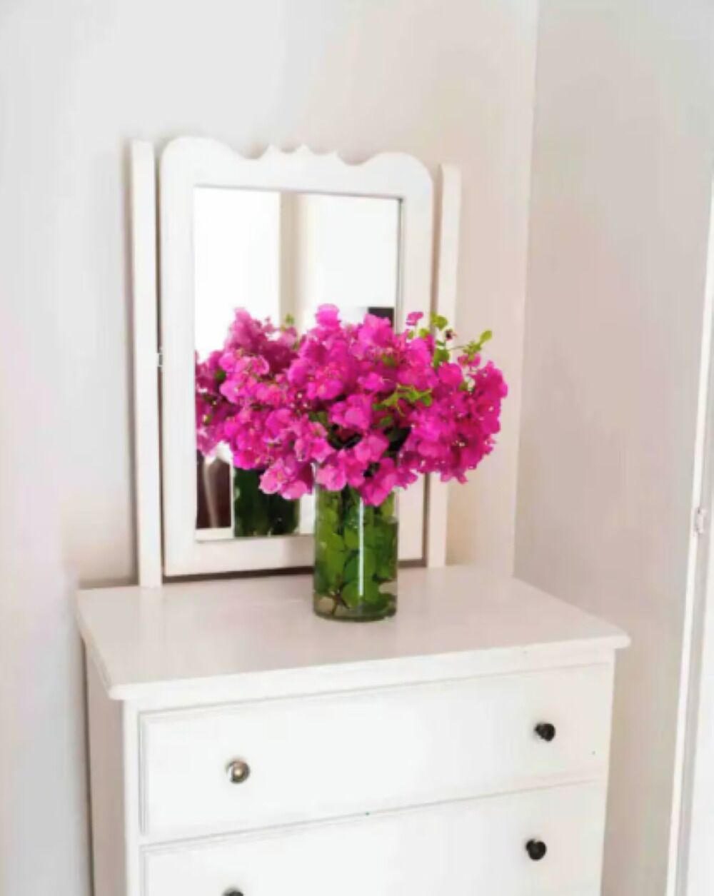 Un aparador blanco con un espejo encima y un florero con flores rosadas en la parte superior.