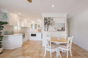 Eine moderne Küche mit weißen Schränken, ein Essbereich mit einem runden Holztisch und weißen Stühlen sowie ein großes Gemälde mit einer Strandansicht.