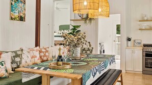 A cozy living area with a sofa, a wooden table with a bottle and glasses, a hanging wicker lamp, and a kitchen in the background.