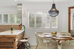 A dining area with a wooden table, wicker chairs, a hanging light fixture, and a window with shutters. - Sandy Daze - Noosa Sound Canalfront Apartment (Noosa Region)