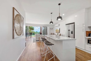 Une cuisine moderne avec des meubles blancs, un îlot central doté de tabourets, des luminaires suspendus et une vue sur l’extérieur.