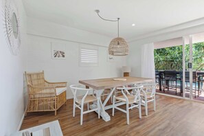 Un espace repas avec une table en bois et des chaises blanches, un luminaire suspendu et une vue sur l’extérieur.