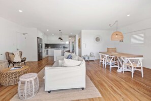 Un salon moderne avec un canapé blanc, des poufs en osier, une table à manger avec des chaises blanches, et une cuisine en arrière-plan.