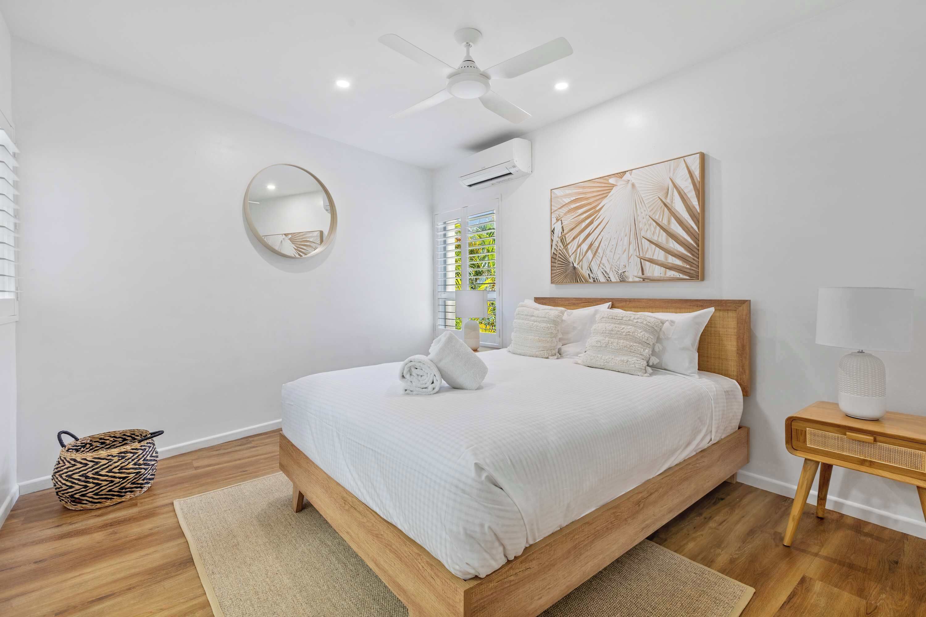 A bedroom with a bed, a ceiling fan, a wall-mounted mirror, a window with blinds, a wooden bedside table with a lamp, and a woven bag.