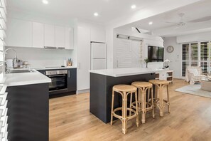A modern kitchen with white cabinetry, a black island, and a dining area with wicker stools. - Modern Noosaville Townhouse - Pool, Tennis, River (Noosa Region)