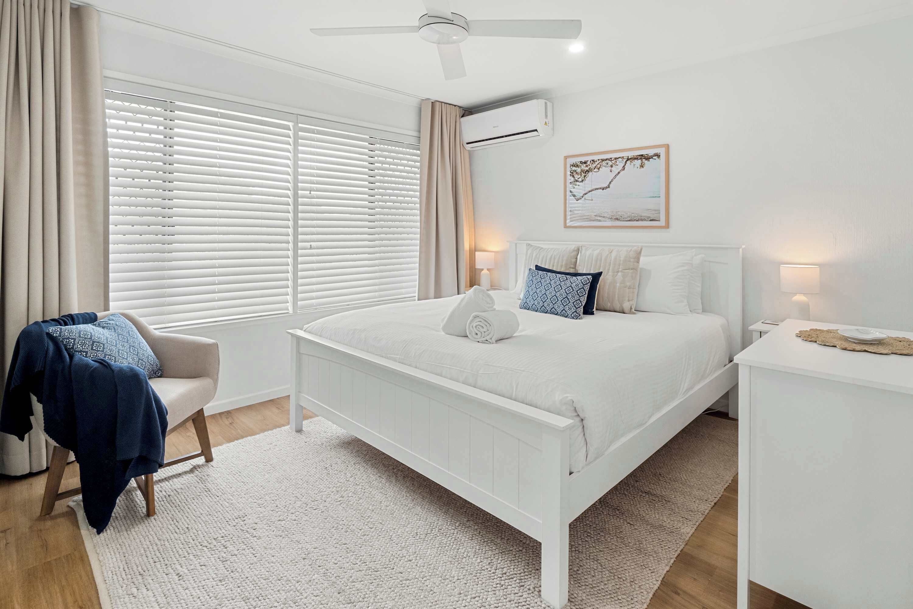 Um quarto com uma cama branca, uma cadeira, uma mesinha de cabeceira, um abajur e um ventilador de teto.