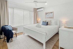 A bedroom with a white bed, a chair, a bedside table, a lamp, and a ceiling fan. - Modern Noosaville Townhouse - Pool, Tennis, River (Noosa Region)